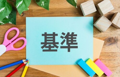 A blue card with the Japanese word for criteria placed on a wooden desk with colored pencils and sticky notes, symbolizing the evaluation standards for mortgage approval