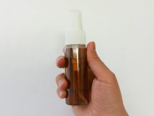 A-hand-holds-a-clear-amber-plastic-spray-bottle-filled-with-a-brown-liquid,-isolated-against-a-white-background